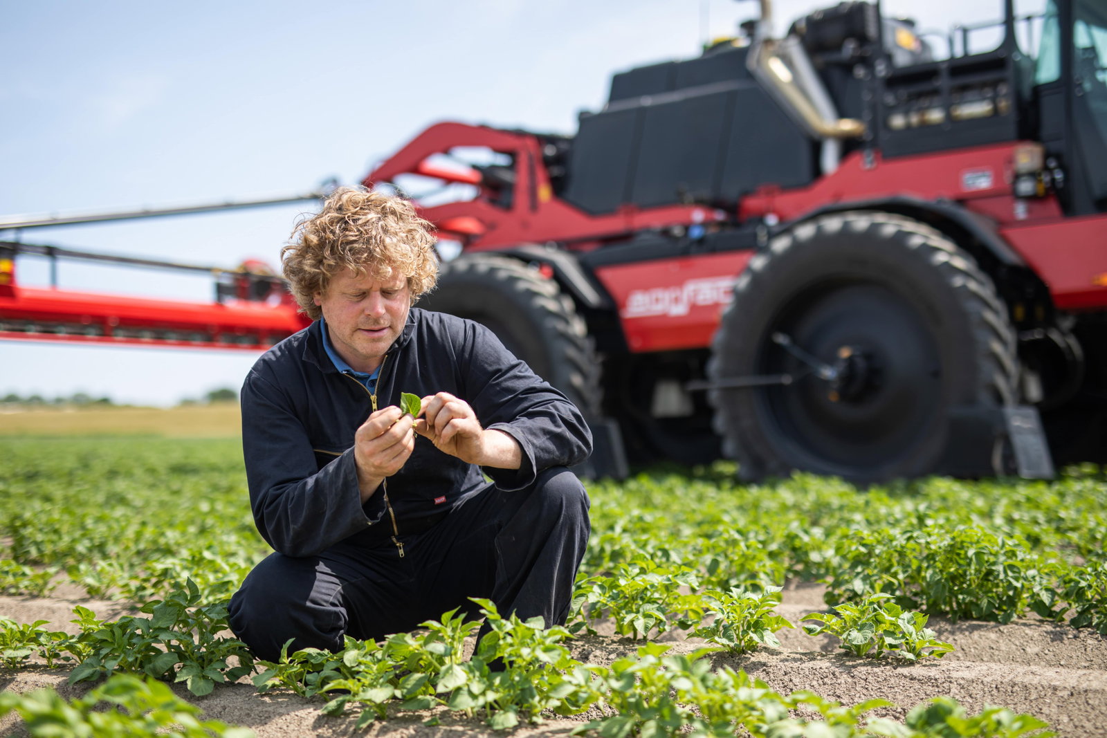 A man is kneeling in a field, closely examining the soil next to a parked Agrifac, symbolizing farming efforts.
