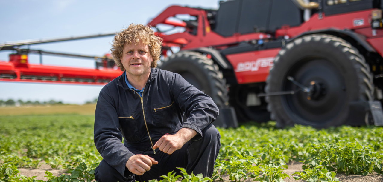 A man kneels in front of an Agrifac sprayer in a field, inspecting the field surrounded by open land and greenery.
