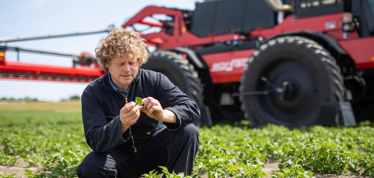 A man is kneeling in a field, closely examining the soil next to a parked Agrifac, symbolizing farming efforts.
