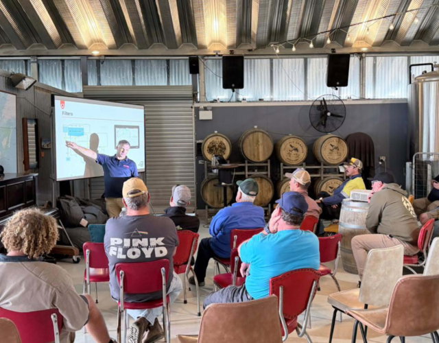 People gathered around a sales representative giving a presentation about Agrifac sprayers at an event.