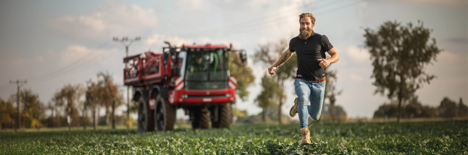 A man runs energetically through a field, with an Agrifac sprayer visible in the background, showcasing rural life and activity.